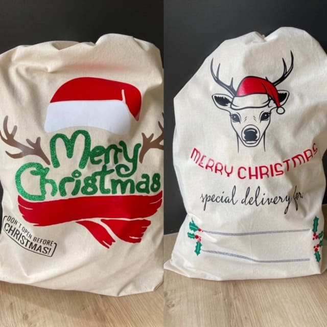 Two personalized Christmas canvas gift sacks by AJ's Infinity Design - left features Santa hat with antlers, sparkly green 'Merry Christmas' text, red scarf accent and 'DON'T OPEN BEFORE CHRISTMAS!' stamp; right shows reindeer wearing Santa hat, red 'MERRY CHRISTMAS' text, 'special delivery for' personalization space with holly decorations, both drawstring holiday bags available in cream canvas