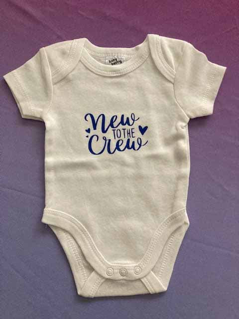 White cotton baby bodysuit with navy blue 'New to the Crew' custom print design featuring decorative heart accent, short-sleeve newborn onesie for baby announcements by AJ's Infinity Design