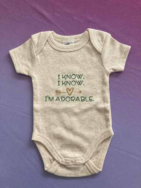 Oatmeal beige baby bodysuit with custom printed funny quote 'I KNOW. I KNOW. I'M ADORABLE.' featuring heart arrow design, 100% cotton short-sleeve infant onesie by AJ's Infinity Design