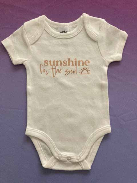 Cream baby bodysuit with 'sunshine for the soul' custom print in gold typography featuring small sun icon, inspirational short-sleeve infant onesie with positive message, soft cotton minimalist design by AJ's Infinity Design