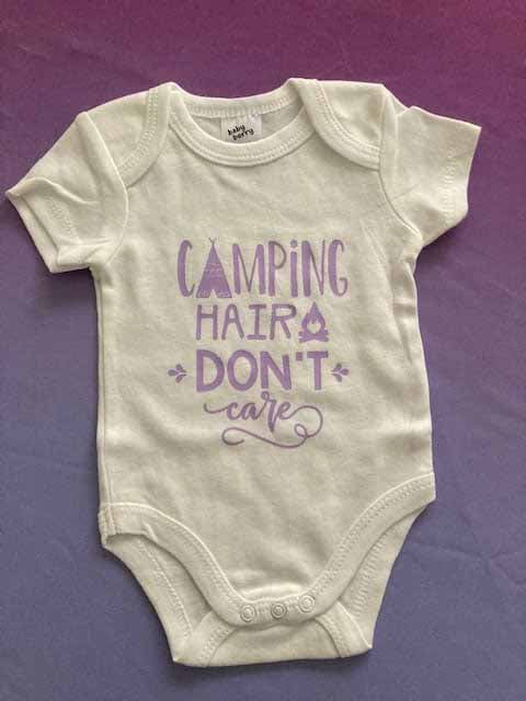 White cotton baby bodysuit with purple 'CAMPING HAIR DON'T CARE' custom print featuring teepee and campfire icons, short-sleeve outdoor adventure themed infant onesie for nature-loving families by AJ's Infinity Design