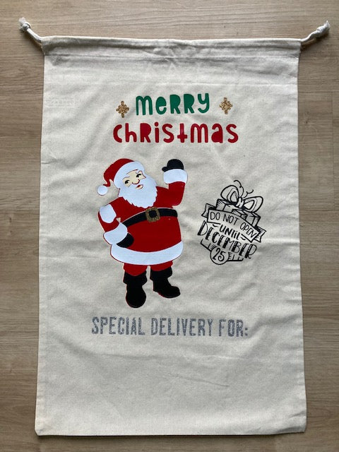 Natural canvas Christmas gift sack with waving Santa Claus illustration, red and green 'MERRY CHRISTMAS' lettering with decorative snowflake accents, 'DO NOT OPEN UNTIL DEC 25' present stamp, and 'SPECIAL DELIVERY FOR:' personalization space, reusable drawstring holiday bag for children's gifts by AJ's Infinity Design