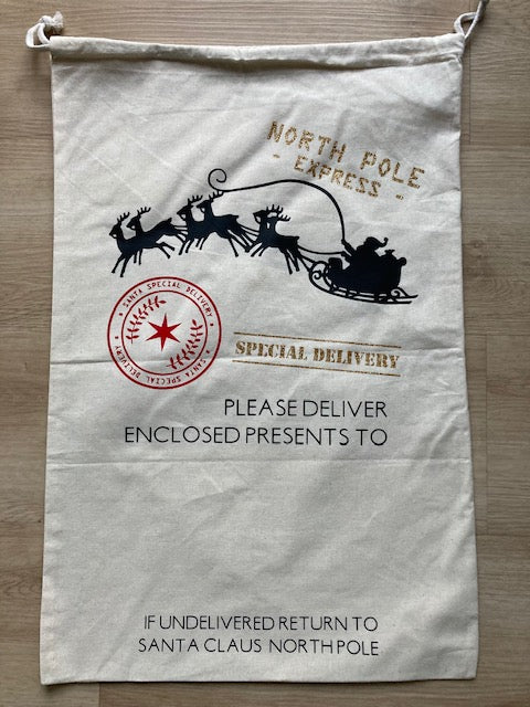 Natural canvas Christmas gift sack with postal theme, featuring gold 'NORTH POLE EXPRESS' heading, black Santa sleigh silhouette, red North Pole official stamp, 'SPECIAL DELIVERY' gold text, personalized delivery address space, and 'IF UNDELIVERED RETURN TO SANTA CLAUS NORTHPOLE' footer, drawstring canvas bag resembling official Santa mail by AJ's Infinity Design
