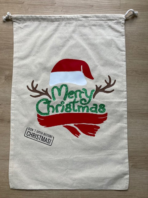 Cream canvas Christmas gift sack featuring Santa hat with reindeer antlers design, sparkly green 'Merry Christmas' text, red scarf accent, 'DON'T OPEN BEFORE CHRISTMAS!' stamp, drawstring closure, reusable holiday present bag for children's gifts by AJ's Infinity Design